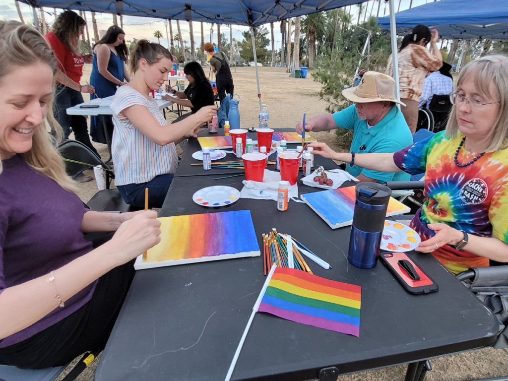 3 women and 1 man sitting at a table in the park painting sunset canvases with the Pride Flag in the foreground