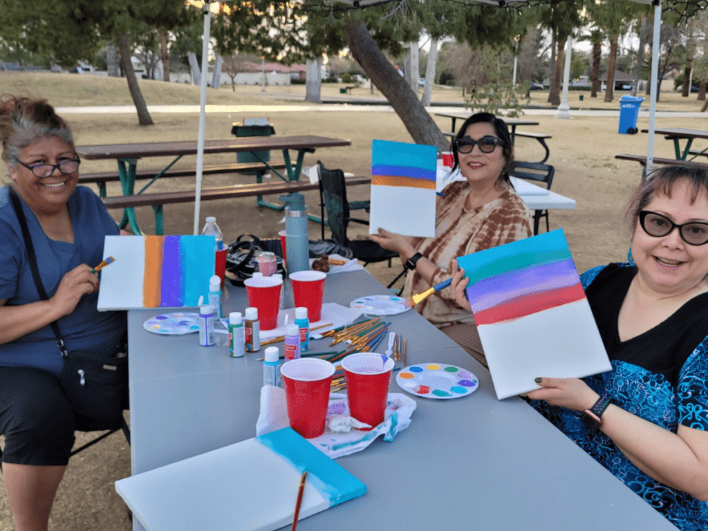 3 women sitting at a table in the park holding up their work in progress paintings