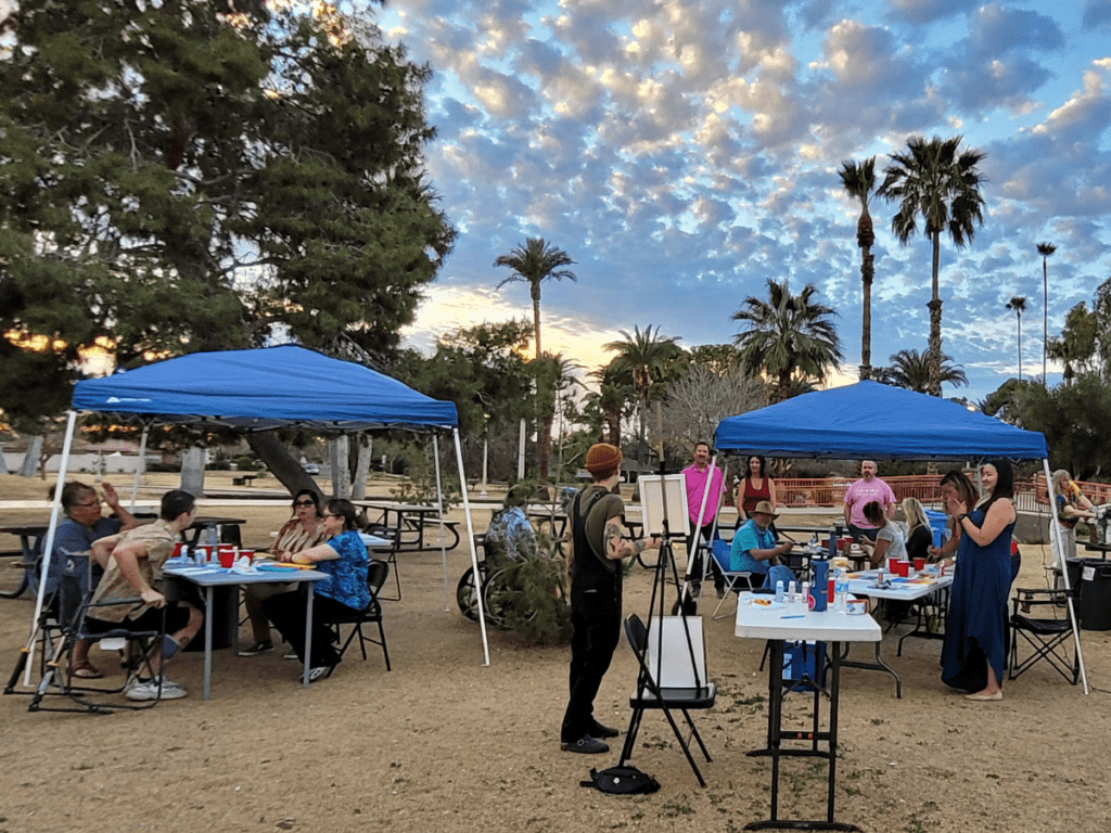 Overview of PHX LGBTQ+ Alliance attendees and teaching artist painting in the park with palm trees and puffy cloud sunset in background