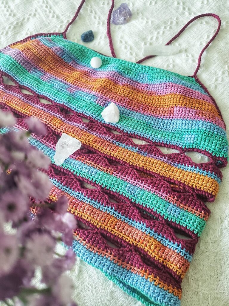 A hand-made crochet top with orange, pink, teal, and red colors laying on a bed next to while and purple crystals.