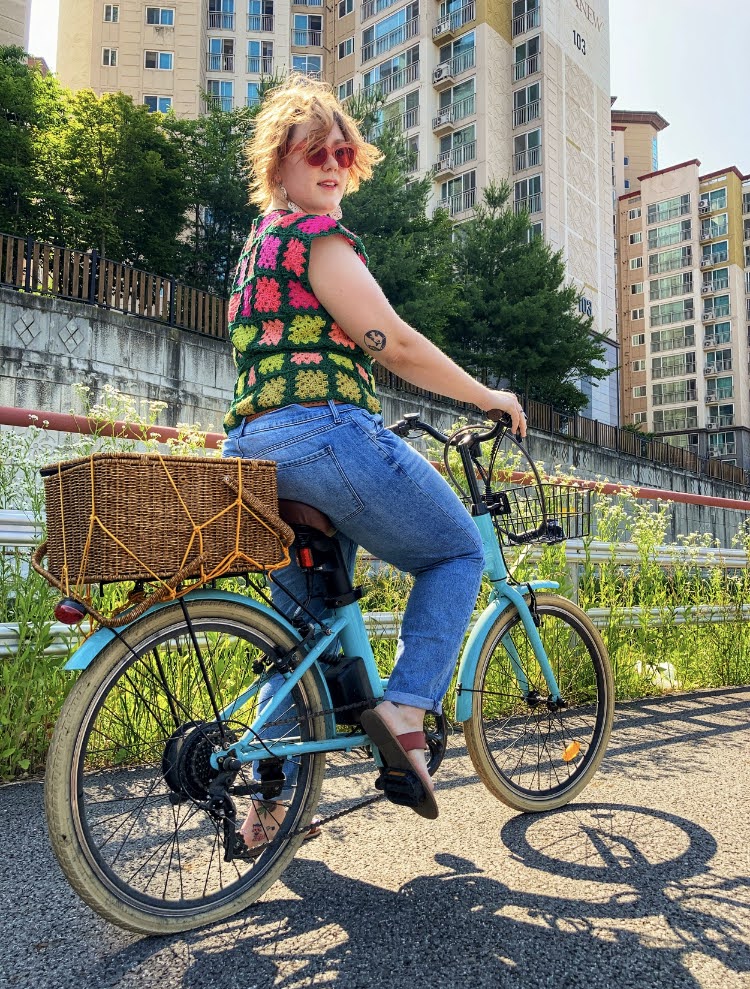 Woman on a bicycle in a city wearing blue jeans, a hand-made pink and green crochet top, and smiling. 