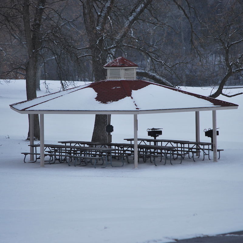A photo of snow covered gazebo with benches and tables underneath it, dead trees framing the background and a blanket of snow covers the ground.  