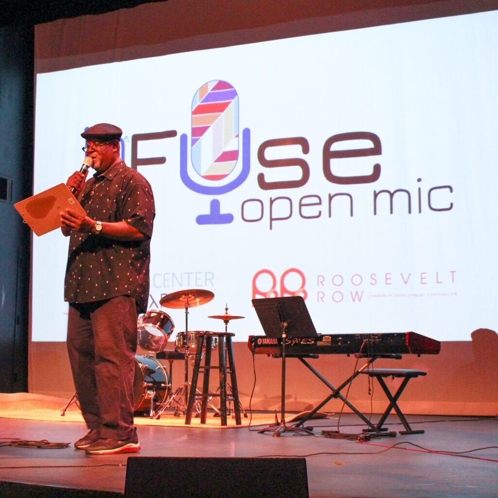 A man wearing a button down shirt and a hat reading from a clipboard on a stage with music equipment behind him